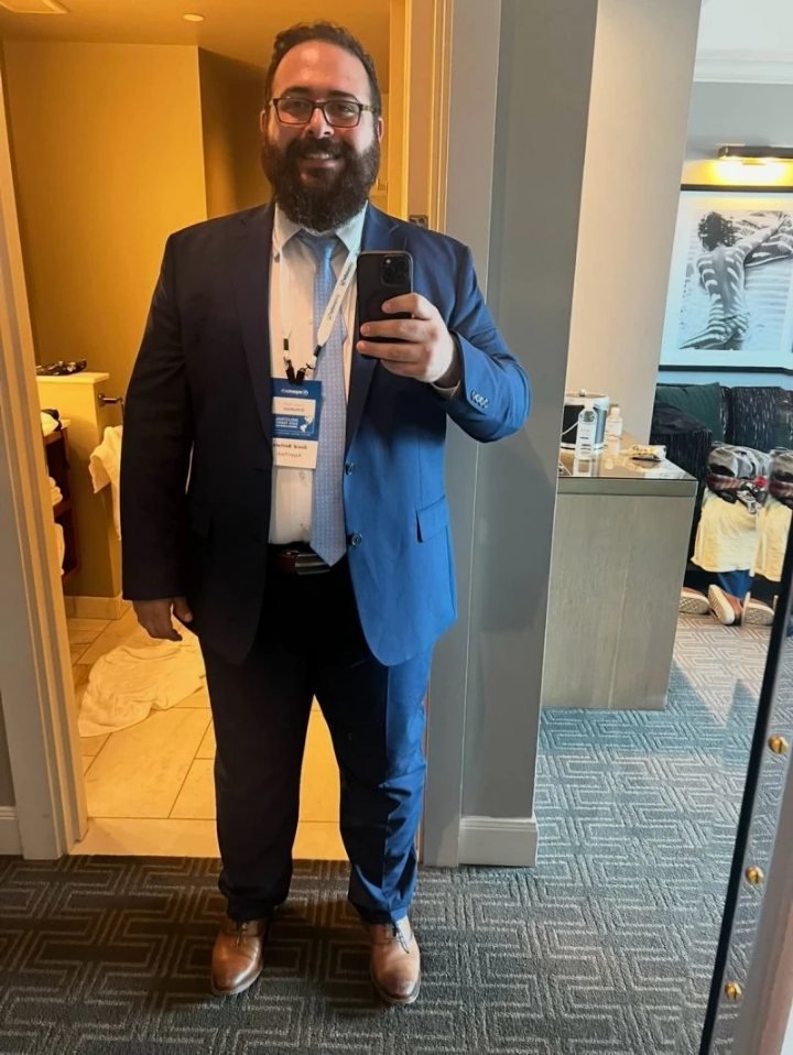 David Bechara in a blue suit and tie with a conference lanyard, taking a mirror selfie in a hotel room.