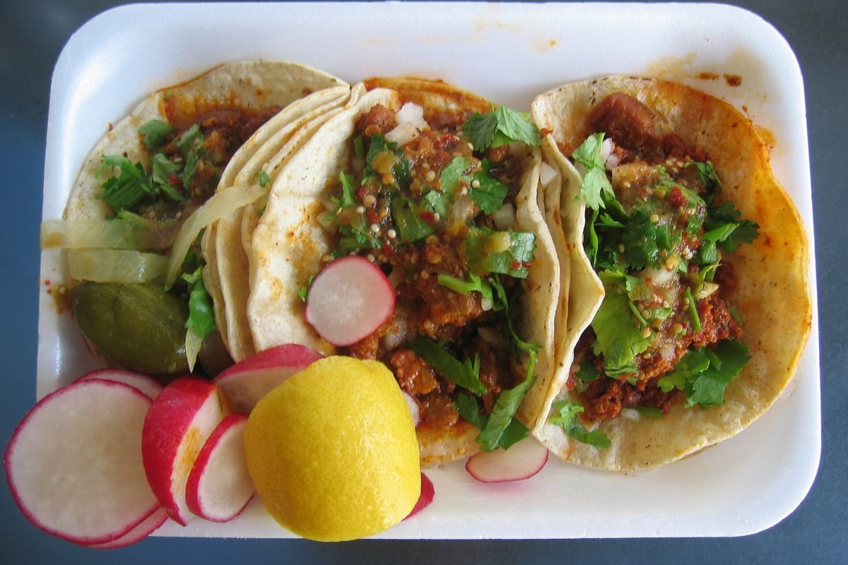 Three soft tacos with meat and cilantro in a foam tray, with sliced radishes and a lemon wedge.