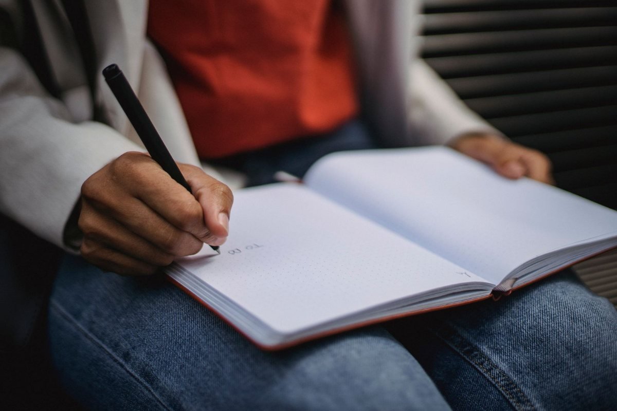 A hand holding a pen over an open notebook resting on a person's lap.