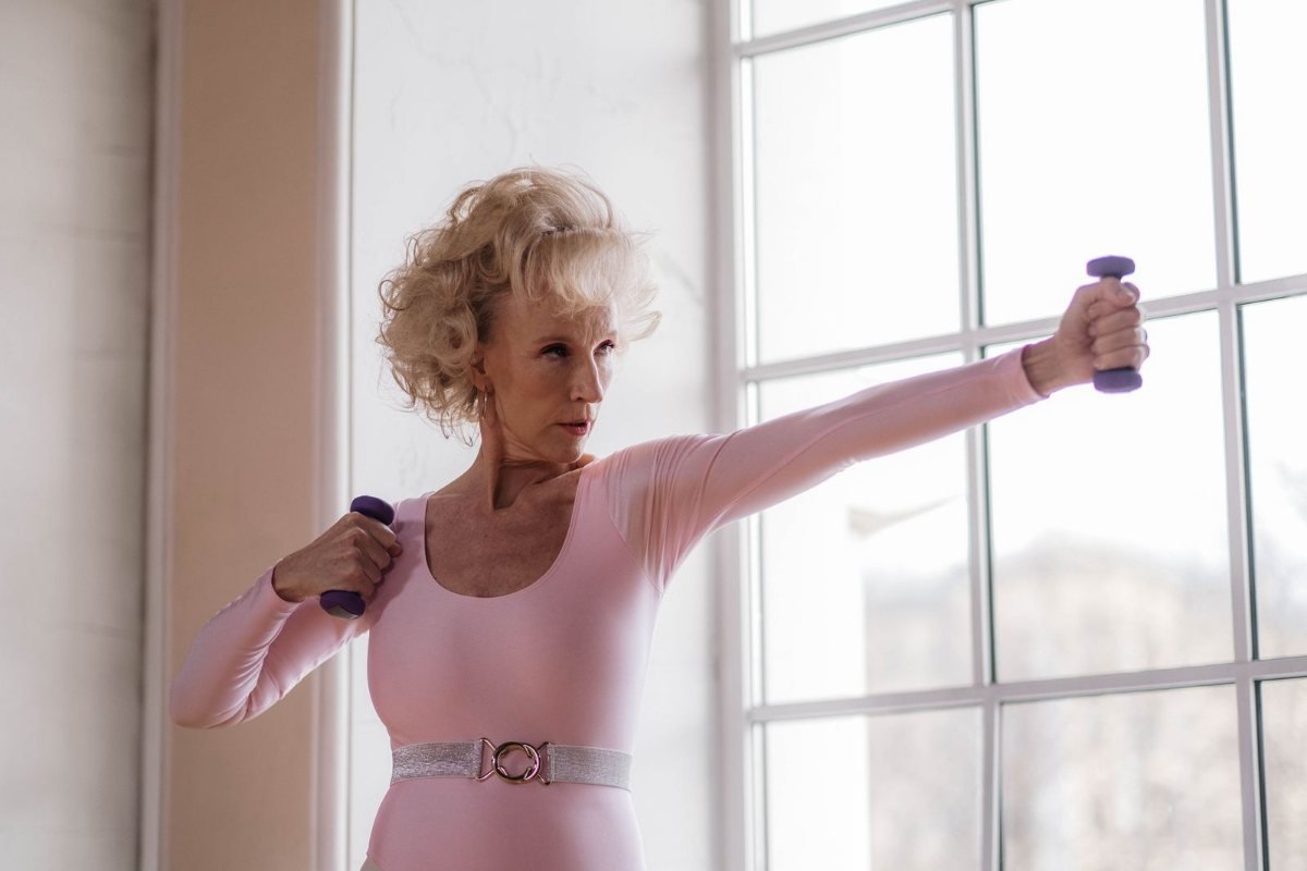 An older woman in a pink top lifting small purple dumbbells beside a large window.