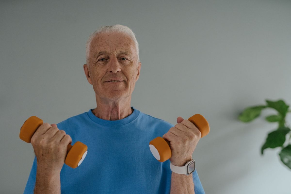An older man in a blue shirt smiling as he lifts two orange dumbbells at home.